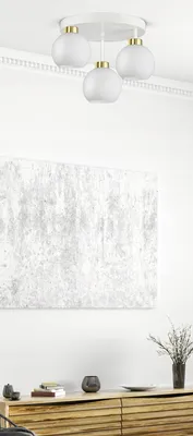 A white ceiling lamp with three glass globes hangs from a white ceiling. Below the lamp, a large abstract painting with a textured gray and white pattern is mounted on a white wall. A wooden cabinet with horizontal slats is visible below the painting, with