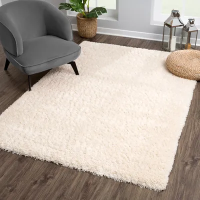 A cream-colored shaggy rug is placed on a dark wooden floor next to a gray armchair. The scene is set in a well-lit room with a large window in the background, showing a plant and decorative lanterns.