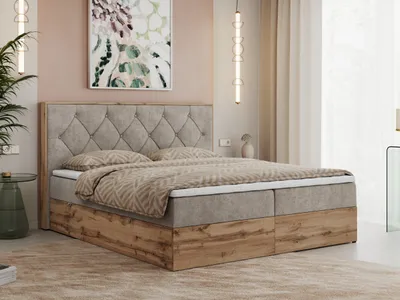 A bedroom scene featuring a large boxspring bed with a tufted headboard and a wooden frame. The bed is made with patterned bedding and is placed in a room with pink walls and beige curtains.