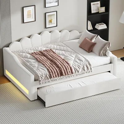 A white daybed with a pull-out trundle bed is shown in a room setting. It has a scalloped headboard and is covered with a quilted blanket and a pink throw blanket.