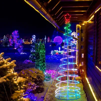A spiral LED Christmas tree decorated with colorful lights, set up outdoors at night with other Christmas decorations in the background.