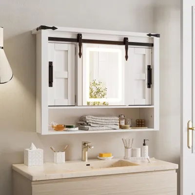 A white medicine cabinet with sliding doors, an illuminated mirror, and an open shelf above a bathroom sink.