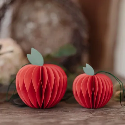 Two red paper apple decorations with green leaves on a wooden surface, with a blurred background.