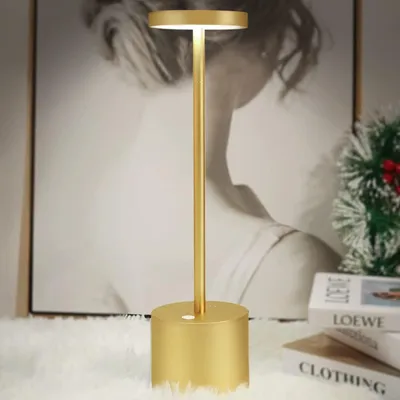 A gold cordless table lamp is displayed on a white fluffy surface in front of a large black and white portrait.