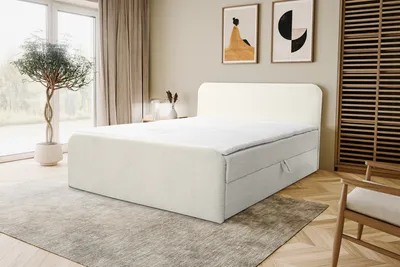 A bedroom scene featuring a beige bouclé fabric boxspring bed with a white headboard, placed on a gray rug in front of a large window with sheer curtains. Two framed abstract art pieces hang on the wall above the bed. A wooden wardrobe is visible on the ri
