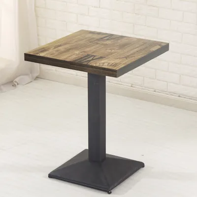 A square-topped table with a dark metal pedestal base and a wood-grain patterned top, set against a white brick wall and white floor.