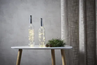 Two glass bottles with fairy lights inside, placed on a white table next to a pine branch and pine cone. A textured curtain is in the background.