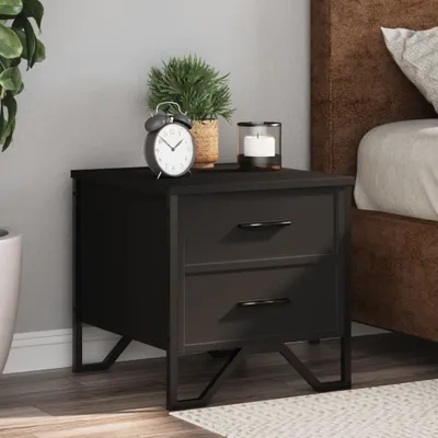 A black nightstand with two drawers is placed next to a bed. An alarm clock, a potted plant, and a candle are on top of the nightstand.