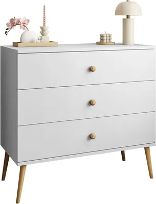 A white dresser with three drawers and wooden legs, decorated with a lamp, candles, and flowers on top.