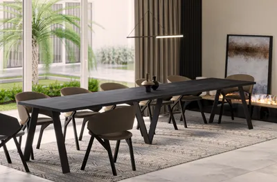A modern dining room with a long black dining table and chairs, set on a patterned rug. Large windows show a palm tree and garden outside. A fireplace with a painting above it is visible in the background.