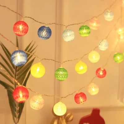A string of colorful cotton ball lights is draped across a wall, with a plant in the foreground.