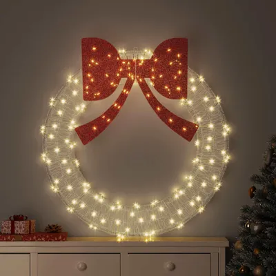 A lighted Christmas wreath with a red bow is displayed on a dresser.