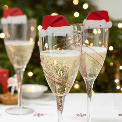 Champagne glasses decorated with Santa hats, set against a blurred Christmas tree background with lights.