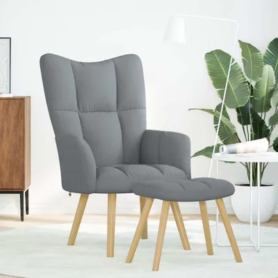 A gray fabric armchair and ottoman are shown in a well-lit room with a large plant and a floor lamp.