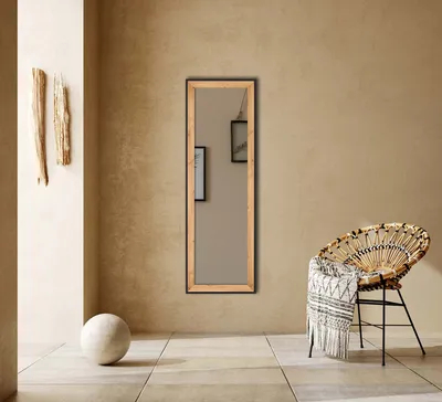 A tall, narrow mirror with a light wood frame stands against a textured tan wall. To the left, two pieces of driftwood are mounted on the wall, and a large, light-colored sphere rests on the floor. To the right, a woven rattan chair with a patterned throw 