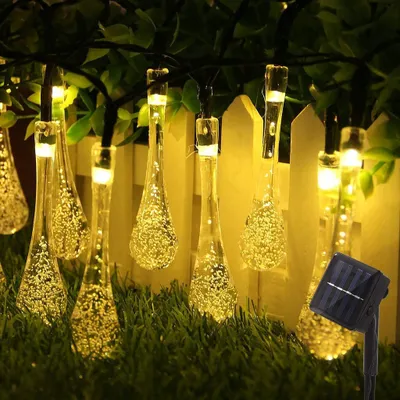 Solar-powered string lights with teardrop-shaped LED bulbs are displayed outdoors, attached to a fence and draped over grass and foliage.