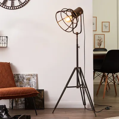 A floor lamp with a cage-like shade is positioned next to an orange corduroy chair and artwork in a room.