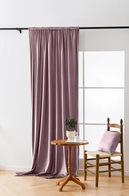 A pink curtain hangs on a rod next to a window. A small wooden table with a plant and a white bottle sits in front of the curtain, and a wooden chair with a pink cushion is to the right of the table.