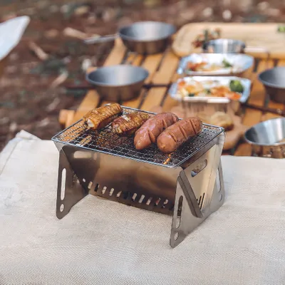 A portable camping grill with sausages cooking on it, set up outdoors on a wooden table.