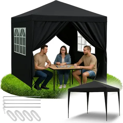 Three people are sitting at a table inside a black gazebo, playing a board game. The gazebo is set up on a grassy area.