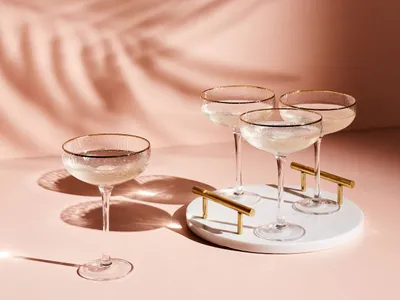 Four champagne glasses with gold rims are arranged on a pink surface, with palm leaf shadows cast on the background.