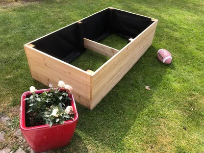 A wooden raised garden bed is shown on a grassy lawn, with a red planter and a football nearby.