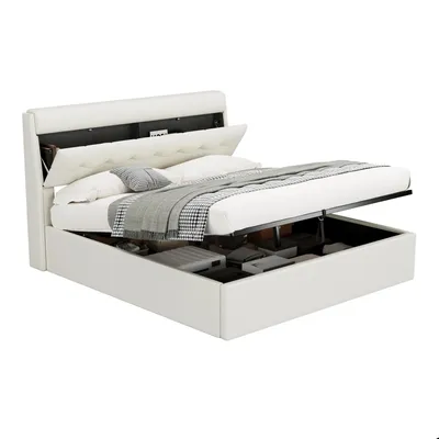 A white upholstered bed frame with its mattress lifted to reveal storage space underneath. The headboard has a hidden compartment. Pillows and a blanket are on the mattress.