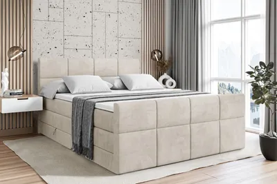 A bedroom scene featuring a large bed with a taupe velvet upholstered frame, white bedding, and a grey throw blanket. The room has a modern aesthetic with textured wall panels, a bedside table, and a large window with curtains.