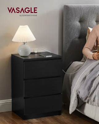 A person is sitting in bed next to a black nightstand with a lamp and phone on it.