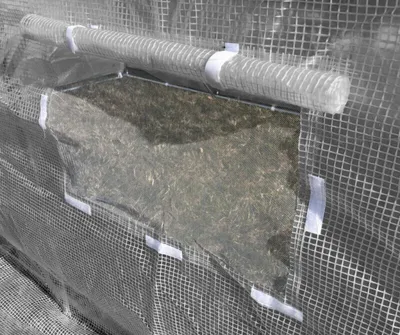 A close-up view of a greenhouse tunnel with a clear plastic window showing soil and vegetation inside. The tunnel is made of a mesh-like material and secured with white straps.