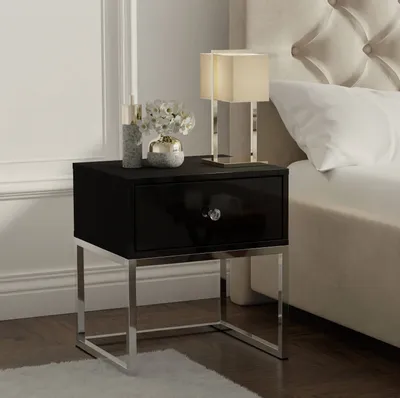 A black bedside table with a silver frame, a drawer with a crystal knob, a lamp, and a vase with flowers. The bedside table is next to a bed with a tufted headboard.
