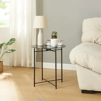 A small round black side table with a glass top is placed next to a beige sofa in a living room. A lamp, a mug, and a small potted plant are on the table.