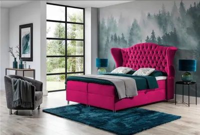 A bedroom scene featuring a large pink tufted bed with teal accents, a gray armchair, and a forest-themed mural on the wall.