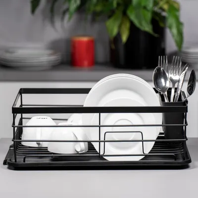 A black metal dish rack filled with white plates and cups, placed on a light gray countertop. In the background, a red cylinder and a green plant in a black pot are visible.