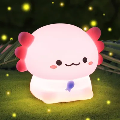 A cute, glowing axolotl-shaped night light sits on a grassy surface with blurred green foliage and yellow bokeh lights in the background.