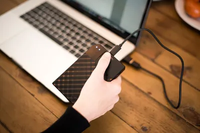 A hand holding an external hard drive connected to a laptop.