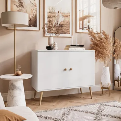 A white two-door cabinet with wooden legs, decorated with a lamp, artwork, and dried flowers in a room.
