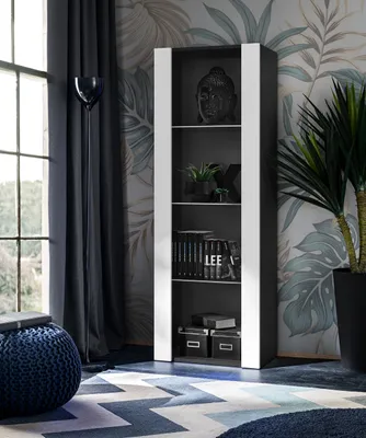 A modern living room with a tall, narrow display cabinet filled with books and decorative items, placed next to a window with dark curtains. A large potted plant stands to the right of the cabinet, and a blue pouf is in the foreground.