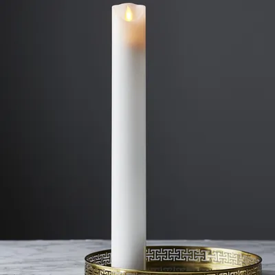 A tall, white LED candle with a flickering yellow flame sits in a decorative gold tray on a marble surface. The background is a dark gray wall.