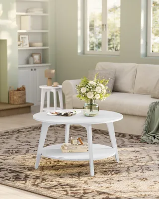 A white round coffee table with a shelf below is placed on a patterned rug in a living room. A vase of flowers sits on the table, and a sofa is visible in the background.