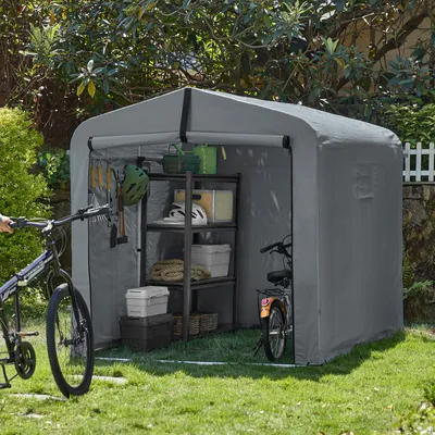 A grey fabric storage shed for bicycles is set up in a grassy yard with trees and a white picket fence in the background. Two bicycles are visible, one being held by a person's hand on the left, and another parked inside the shed on the right. Shelving uni