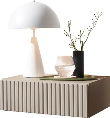 A white table lamp, a black vase with a plant, and a glass of water are placed on a ribbed gray nightstand.