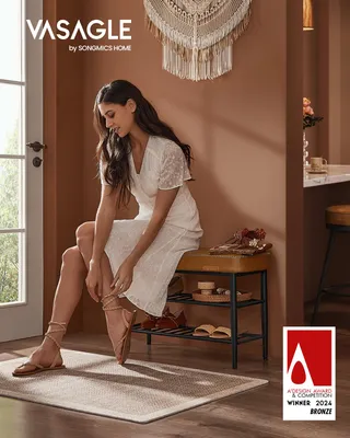 A woman is sitting on a shoe bench in an entryway, adjusting her sandal. The bench has a brown upholstered top and black metal shelves holding shoes and decorative items. A macrame wall hanging is visible on the wall.