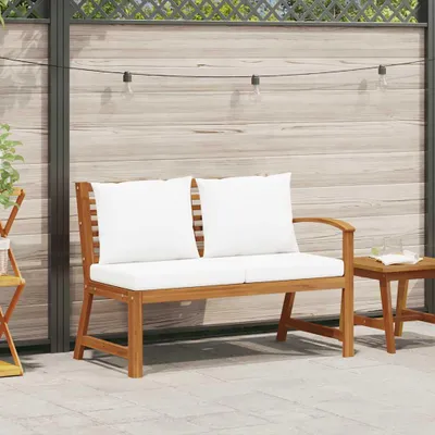 A wooden garden bench with white cushions is placed on a paved patio in front of a wooden fence.