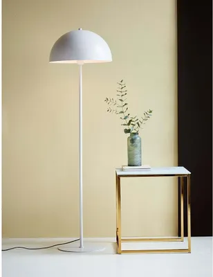 A white floor lamp stands next to a small gold-framed side table with a vase of greenery.
