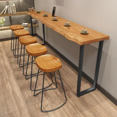 A long, narrow wooden bar table with a live edge, paired with several wooden stools, set against a light gray wall and wooden floor.