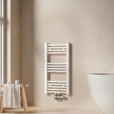 A white towel radiator is mounted on a light beige wall in a bathroom setting, next to a wooden stool with a folded towel.