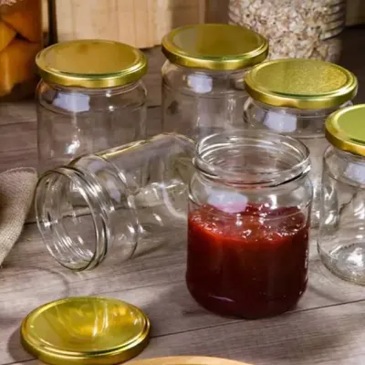 Several glass jars with gold lids are arranged on a wooden surface. One jar in the foreground contains a red jam or preserve.