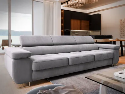 A modern living room with a large grey sofa in the foreground, a kitchen area in the background, and a large window with a view of water and hills.