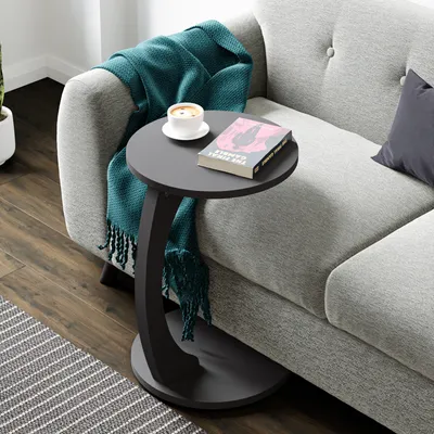 A dark gray C-shaped side table with a book and a cup of coffee on it, placed next to a gray sofa with a teal throw blanket.